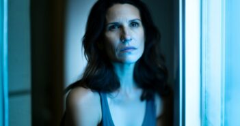 A woman with wavy brown hair sits by a window, illuminated by cool blue light, looking slightly upward with a serious, contemplative expression. She wears a sleeveless top and is framed by soft-focus surroundings.
