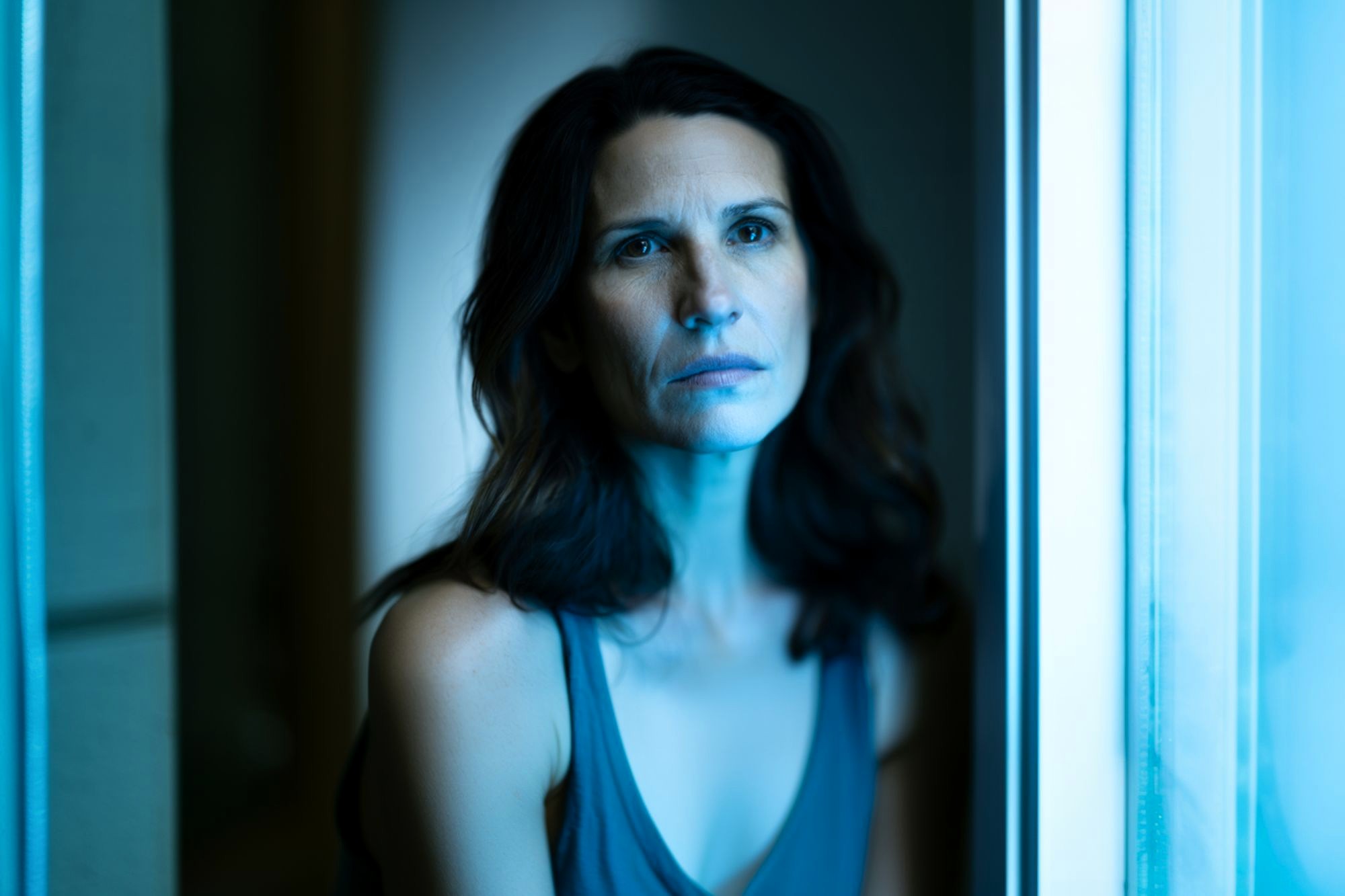 A woman with wavy brown hair sits by a window, illuminated by cool blue light, looking slightly upward with a serious, contemplative expression. She wears a sleeveless top and is framed by soft-focus surroundings.
