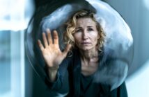 A woman with curly blonde hair touches the inside of a transparent bubble, looking out with a serious, contemplative expression. The background is softly blurred.