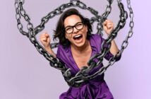 A woman in glasses and a purple shirt smiles widely with clenched fists, surrounded by large metal chains against a light purple background.