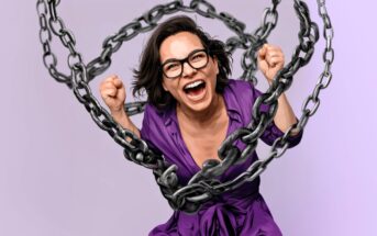 A woman in glasses and a purple shirt smiles widely with clenched fists, surrounded by large metal chains against a light purple background.