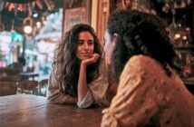 Two people sit across from each other at a wooden table in a warmly lit cafe. One person smiles, resting their chin on their hand, while listening to the other. Cozy, colorful decorations are visible in the background.