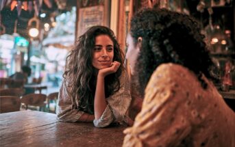 Two people sit across from each other at a wooden table in a warmly lit cafe. One person smiles, resting their chin on their hand, while listening to the other. Cozy, colorful decorations are visible in the background.