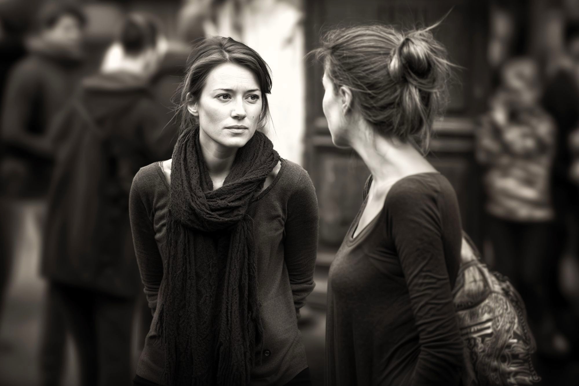 Two women stand facing each other, engaged in conversation on a busy street. One looks serious, while the other is turned slightly away. The background is blurred with people walking. The image is in black and white.
