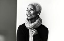 A stylish older woman with short gray hair smiles while looking to the side. She wears a textured scarf and a dark sweater, standing against a bright, minimalist background. The image is black and white.