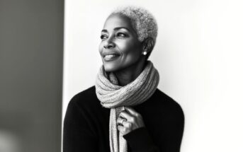 A stylish older woman with short gray hair smiles while looking to the side. She wears a textured scarf and a dark sweater, standing against a bright, minimalist background. The image is black and white.
