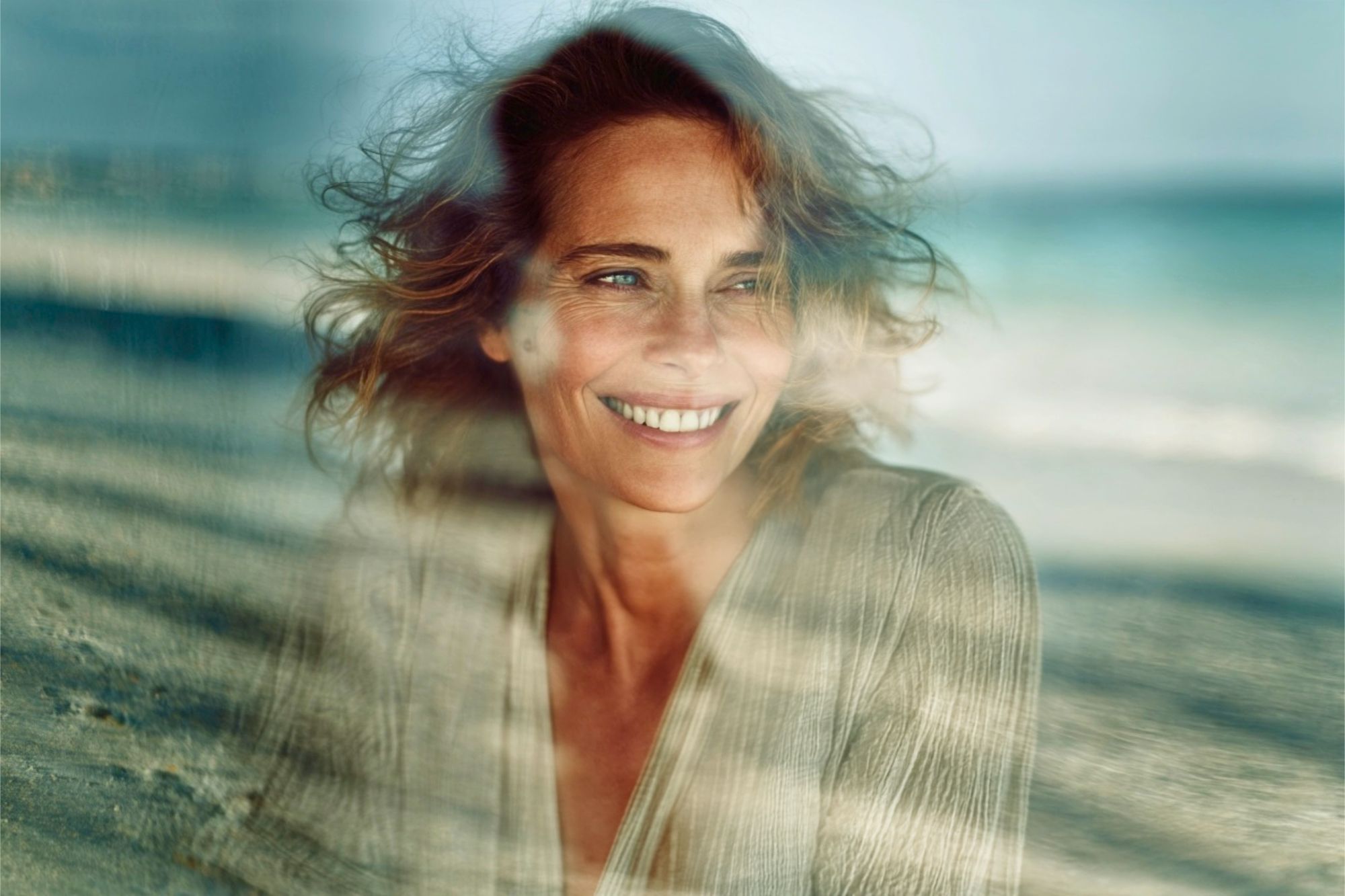 A smiling woman with light brown hair viewed through a soft, double-exposed effect overlaying a beach scene.