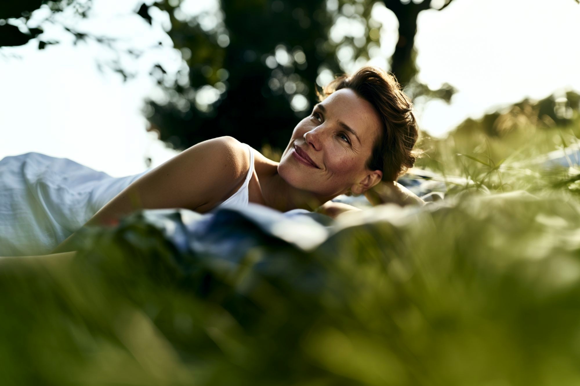 A woman in a white dress is lying on a blanket outdoors, smiling softly and looking into the distance, surrounded by green grass and trees in natural sunlight.