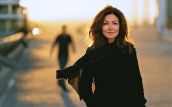 A woman in a black outfit stands in the foreground while a blurred figure walks behind her in the distance at sunset.