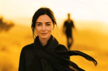 A woman with dark hair and a black scarf stands in focus against a bright yellow background, while a blurry figure walks in the distance behind her.