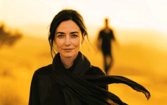 A woman with dark hair and a black scarf stands in focus against a bright yellow background, while a blurry figure walks in the distance behind her.