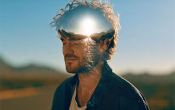 A double exposure portrait of a man with his eyes closed, featuring a bright sun setting over mountains superimposed onto his head.