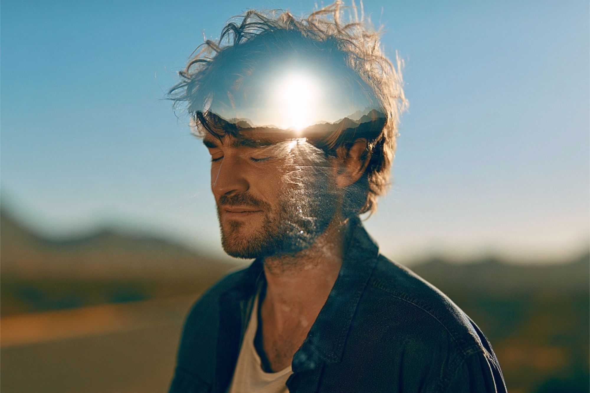 A double exposure portrait of a man with his eyes closed, featuring a bright sun setting over mountains superimposed onto his head.