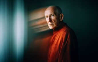 An older man in a red shirt stands by a window, looking serious. The background is dark, and there's a blurred double-exposure effect, creating a ghostly duplicate of his face to the left.