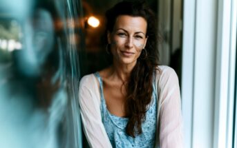 A woman with long brown hair, hoop earrings, and face paint smiles softly while leaning against a reflective glass surface, wearing a blue top and light cardigan.