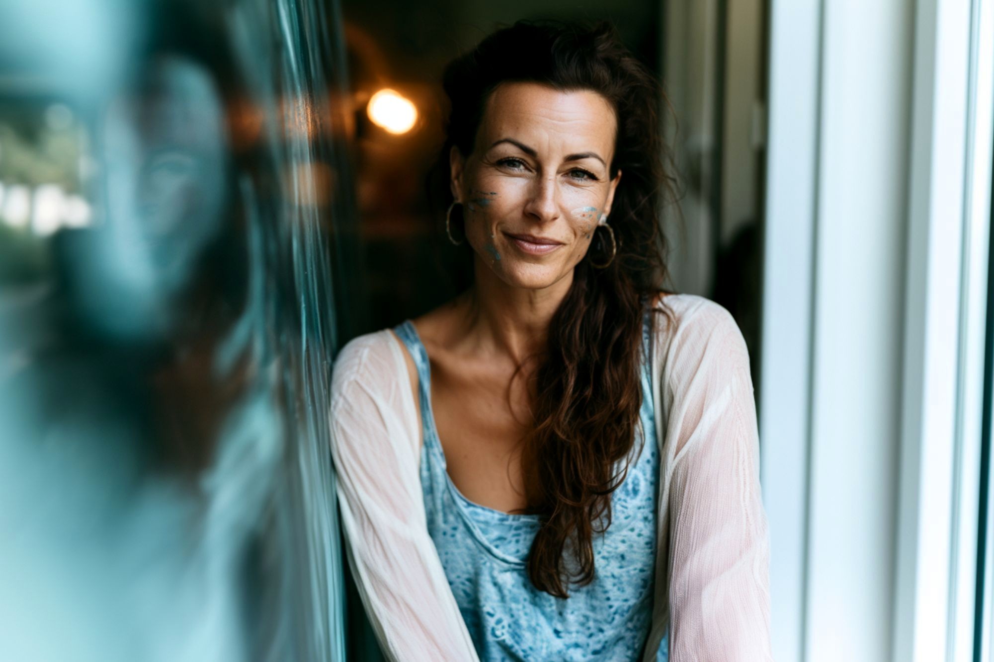 A woman with long brown hair, hoop earrings, and face paint smiles softly while leaning against a reflective glass surface, wearing a blue top and light cardigan.