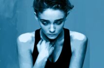 A stylized blue-toned portrait of a woman looking down, appearing thoughtful or sad, with her hand near her neck and wearing a sleeveless black top against a plain background.