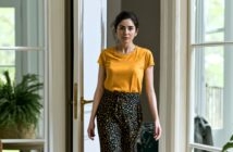 A woman with dark hair in a ponytail, wearing a yellow T-shirt and patterned pants, stands indoors near a glass door with potted plants visible in the background.