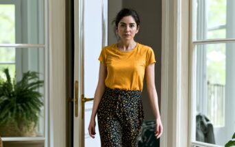 A woman with dark hair in a ponytail, wearing a yellow T-shirt and patterned pants, stands indoors near a glass door with potted plants visible in the background.
