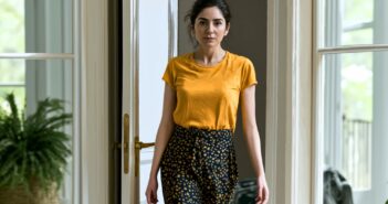 A woman with dark hair in a ponytail, wearing a yellow T-shirt and patterned pants, stands indoors near a glass door with potted plants visible in the background.