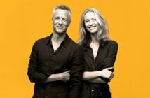 A grayscale photo of a man and a woman standing side by side, both smiling and wearing dark shirts, with a bright yellow background.