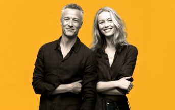 A grayscale photo of a man and a woman standing side by side, both smiling and wearing dark shirts, with a bright yellow background.