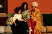Three generations of women sit on a couch: a young girl is crying, a middle-aged woman looks somber, and an elderly woman in glasses gently comforts them, against a warm orange background.