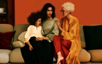 Three generations of women sit on a couch: a young girl is crying, a middle-aged woman looks somber, and an elderly woman in glasses gently comforts them, against a warm orange background.