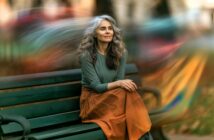 A woman with long gray hair sits on a park bench, wearing a green top and orange skirt. She looks peaceful, with blurred colorful lights swirling in the background, creating a dreamy atmosphere.