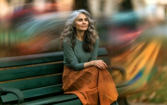 A woman with long gray hair sits on a park bench, wearing a green top and orange skirt. She looks peaceful, with blurred colorful lights swirling in the background, creating a dreamy atmosphere.