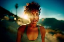 A woman with curly hair stands outdoors in the sunlight, wearing a tank top. The background is blurred, showing a mountainous landscape and a bright, glowing sun behind her.