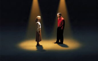 A woman and a man stand facing each other, each illuminated by their own individual spotlight against a dark background.