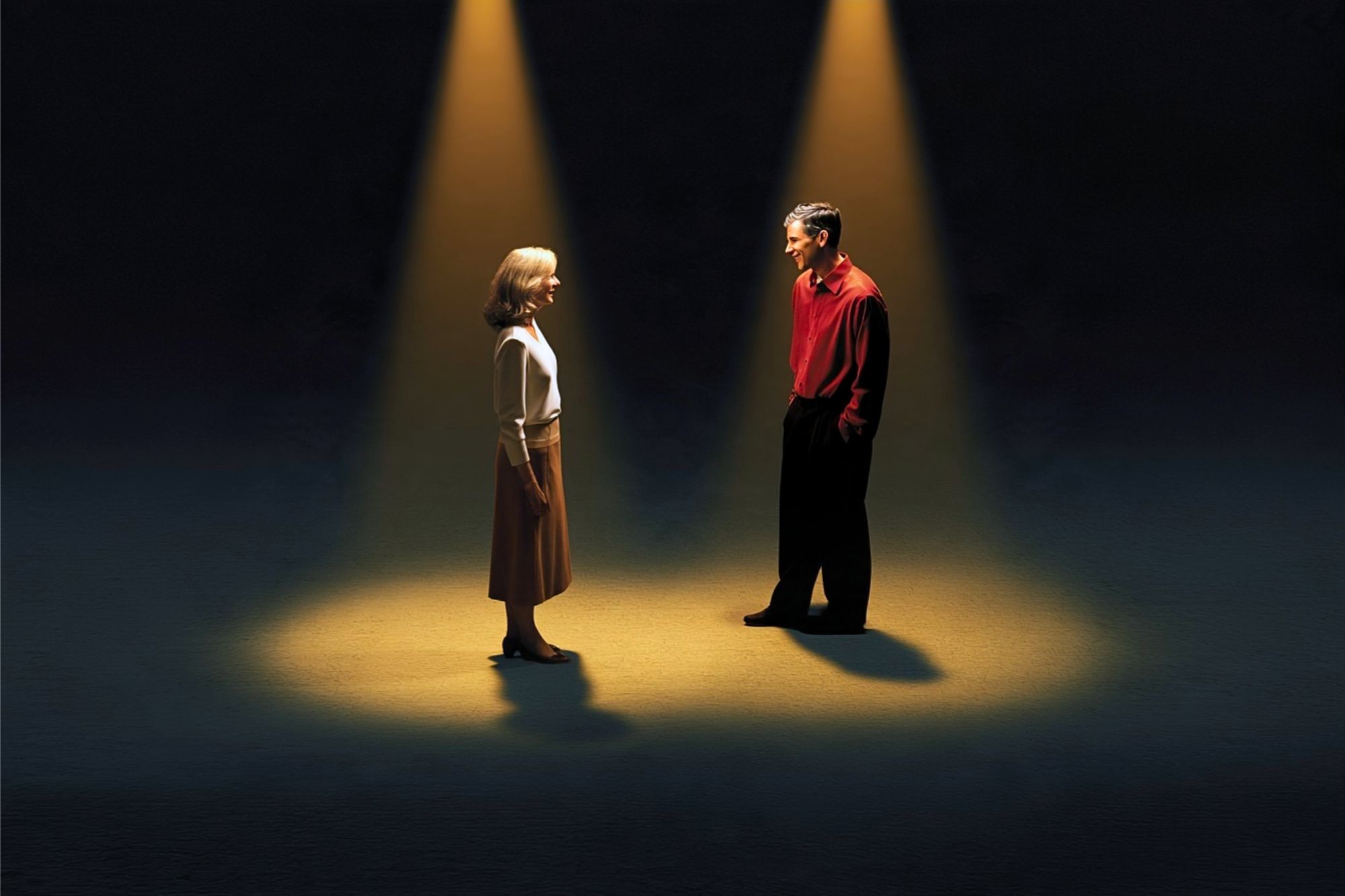 A woman and a man stand facing each other, each illuminated by their own individual spotlight against a dark background.