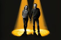 A smiling woman and man holding hands stand together under two bright yellow spotlights, surrounded by darkness.