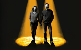 A smiling woman and man holding hands stand together under two bright yellow spotlights, surrounded by darkness.