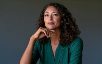 A woman with long, curly dark hair wearing a green top, resting her chin on her hand while looking upward.