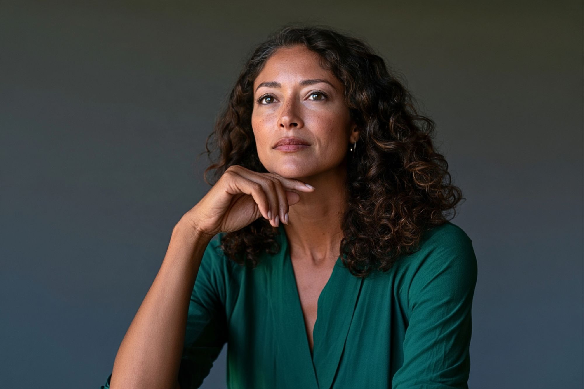 A woman with long, curly dark hair wearing a green top, resting her chin on her hand while looking upward.