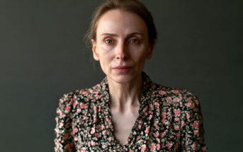 A woman with a neutral expression wearing a black floral-print blouse poses against a dark background.