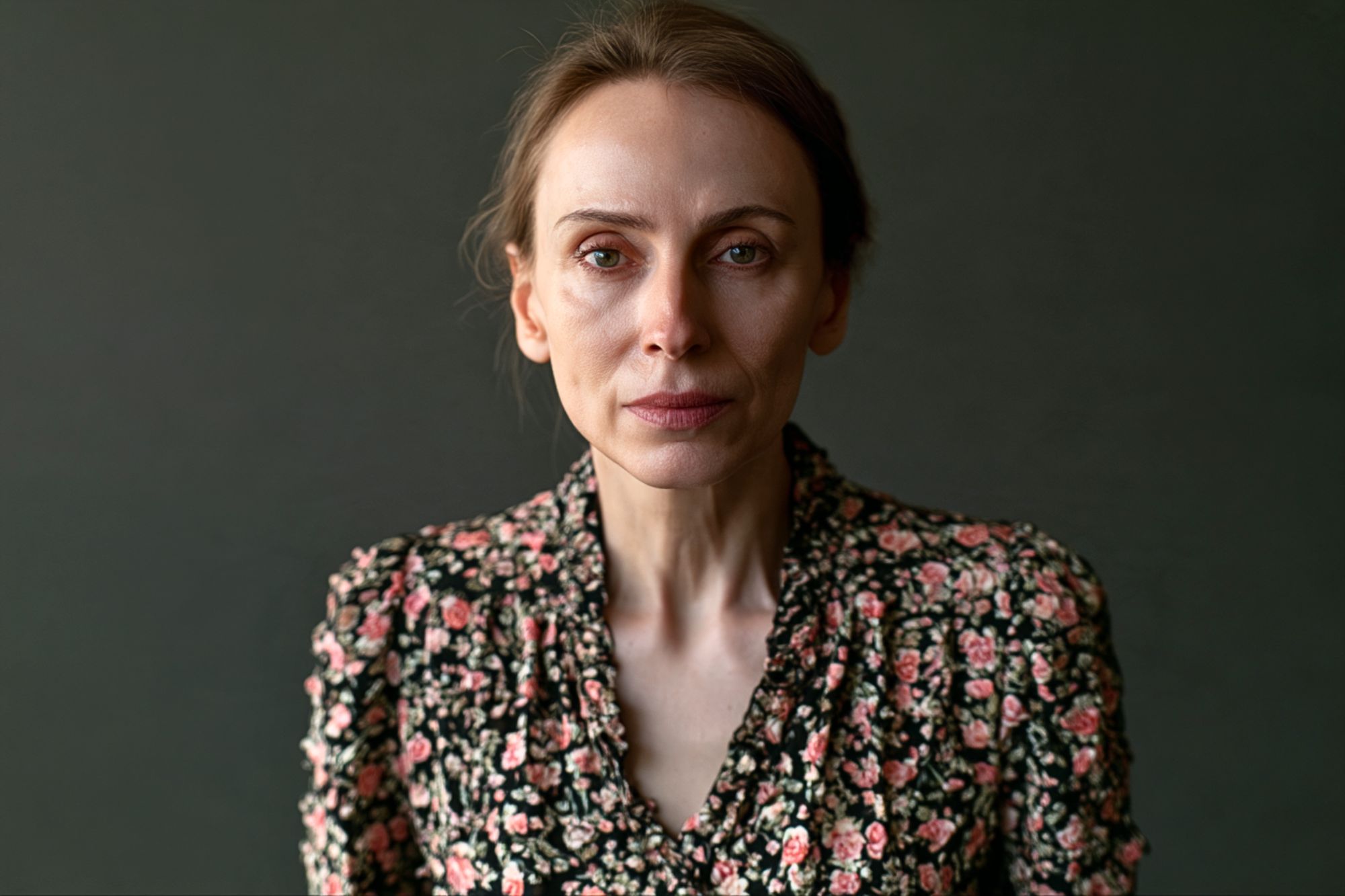 A woman with a neutral expression wearing a black floral-print blouse poses against a dark background.