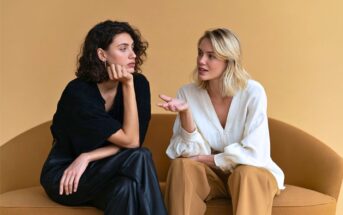 Two women sit on a tan couch against a light brown background. One woman gestures while speaking, and the other listens with her chin resting on her hand. Both have serious expressions and casual, stylish clothing.