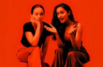 Two women sit side by side against a red background; one looks bored with her chin on her hand, while the other gestures animatedly, appearing to tell an engaging story.