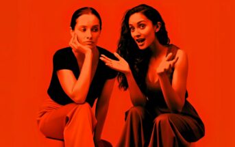 Two women sit side by side against a red background; one looks bored with her chin on her hand, while the other gestures animatedly, appearing to tell an engaging story.