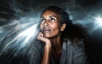 A woman with curly hair gazes upward thoughtfully, resting her chin on her hand, surrounded by dramatic, glowing light effects in the background.