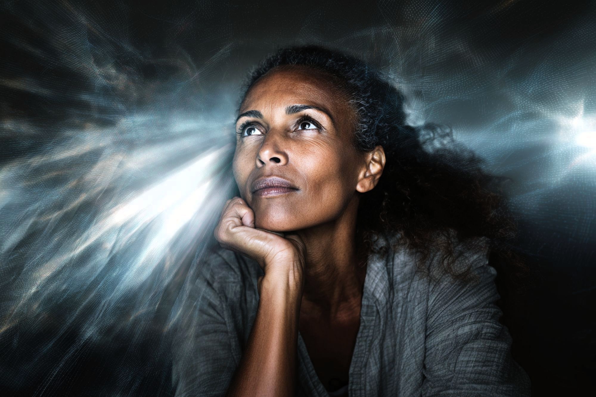 A woman with curly hair gazes upward thoughtfully, resting her chin on her hand, surrounded by dramatic, glowing light effects in the background.