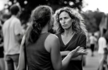 Two women stand outside, facing each other in conversation. One woman has curly hair and crosses her arms, looking serious, while the other gestures with her hand. There are blurred people in the background.