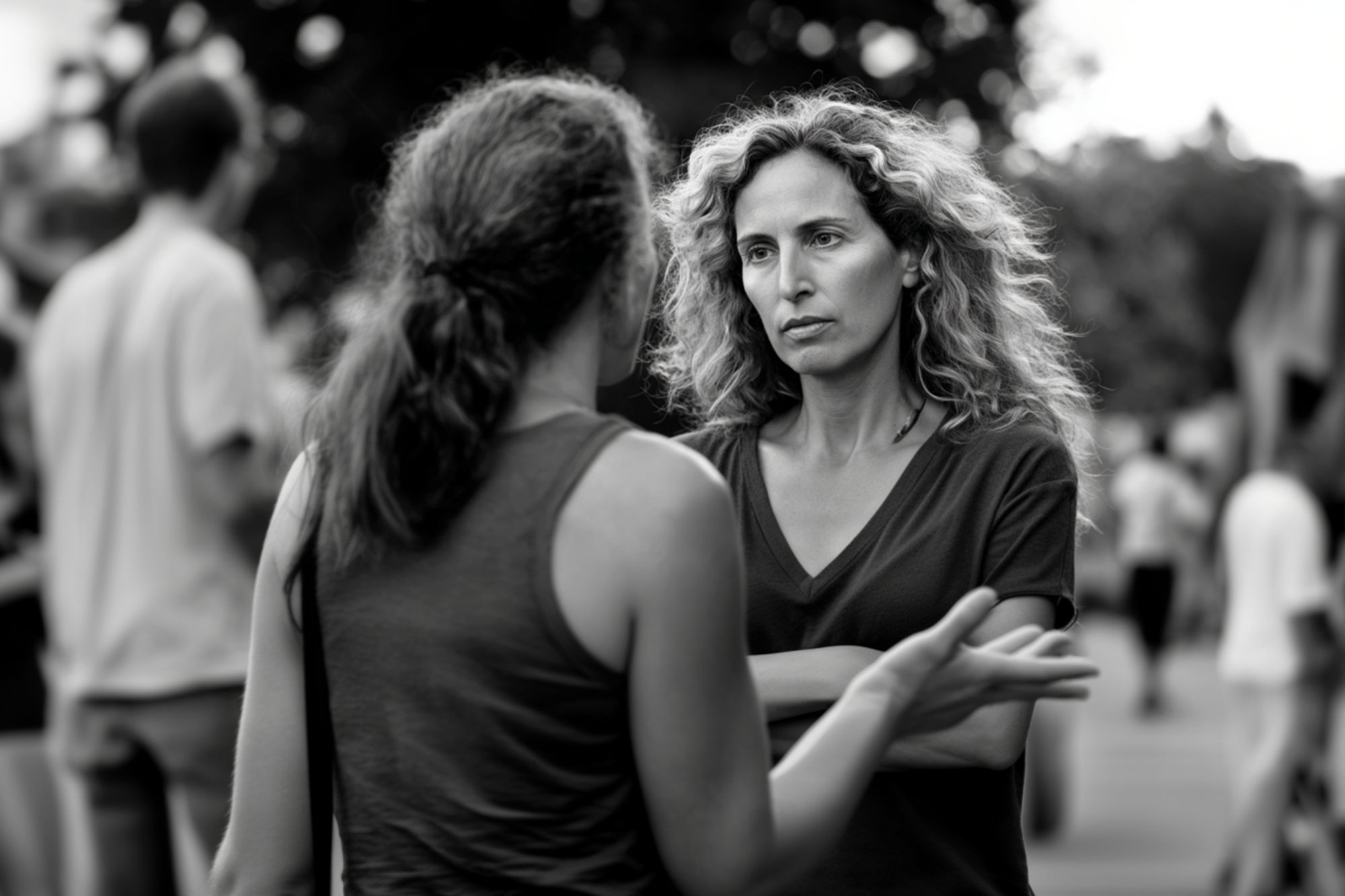 Two women stand outside, facing each other in conversation. One woman has curly hair and crosses her arms, looking serious, while the other gestures with her hand. There are blurred people in the background.