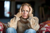 A woman in a tan sweater sits indoors, holding her head with both hands and looking forward with a serious, worried expression.