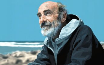 An older man with a beard and balding head, wearing a hooded jacket, sits by the beach gazing thoughtfully into the distance under a clear blue sky.