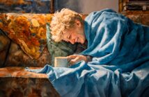 A smiling person wrapped in a blue blanket sits on a floral couch, holding a mug. Warm sunlight lights up their face, and a yellow-patterned pillow is nearby. The scene feels cozy and relaxed.