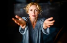 A blonde woman in a gray top extends her arms forward with an expressive, emotional look on her face against a dark blurred background.